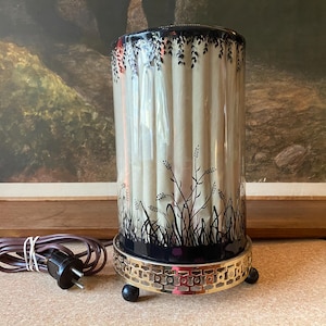 Puede incluir: Una lámpara cilíndrica con base negra y dorada, con un diseño floral en blanco y negro. La lámpara tiene un interior color crema y una parte superior negra. Un cable eléctrico marrón con enchufe está al lado de la lámpara.
