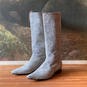 May include: A pair of knee-high boots with a light gray and black leopard print pattern. The boots have a pointed toe and a low heel. The background features a vintage landscape painting.