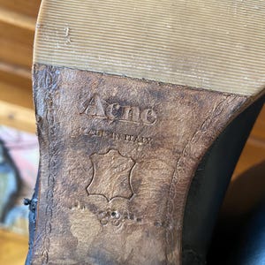 May include: Close-up of the sole of a black leather boot. The brand name "Acne" is embossed on the brown leather sole, with "MADE IN ITALY" below it. The sole is a light tan color.