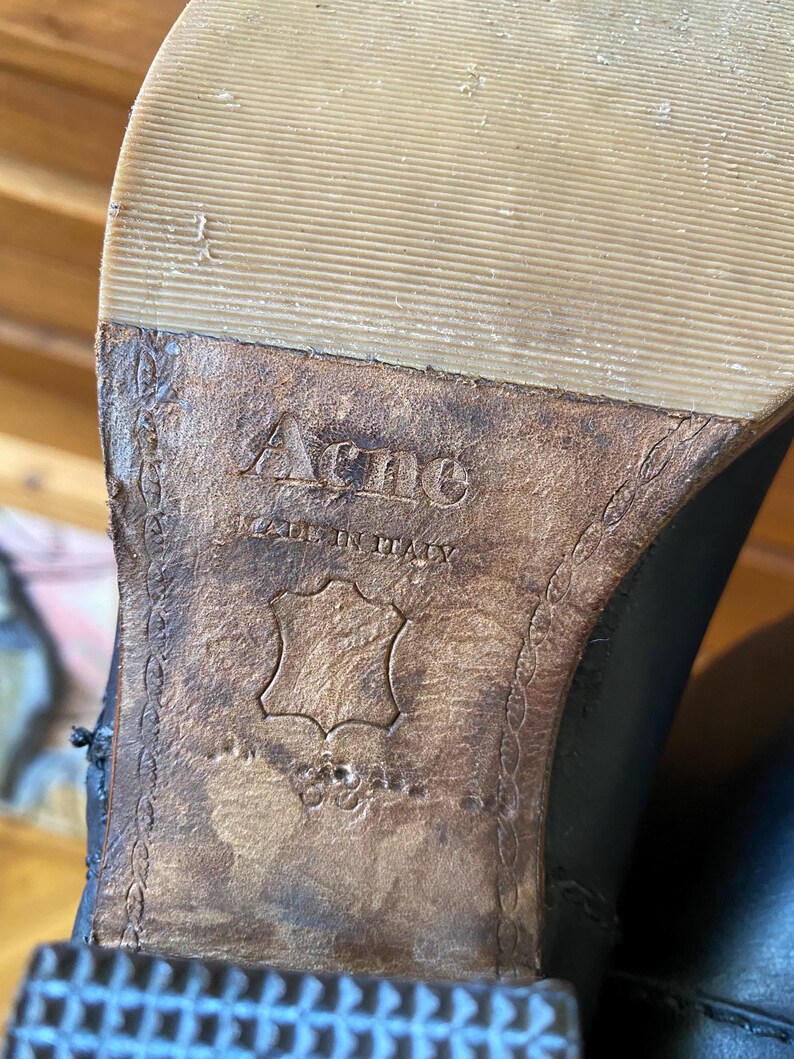 May include: Close-up of the sole of a black leather boot. The brand name "Acne" is embossed on the brown leather sole, with "MADE IN ITALY" below it. The sole is a light tan color.