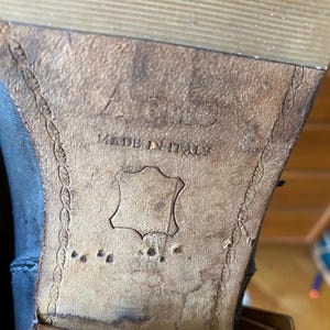 May include: Close-up of the sole of a black leather boot. The sole is made of brown leather and features the text "ACIO MADE IN ITALY". The leather is worn and shows signs of use.