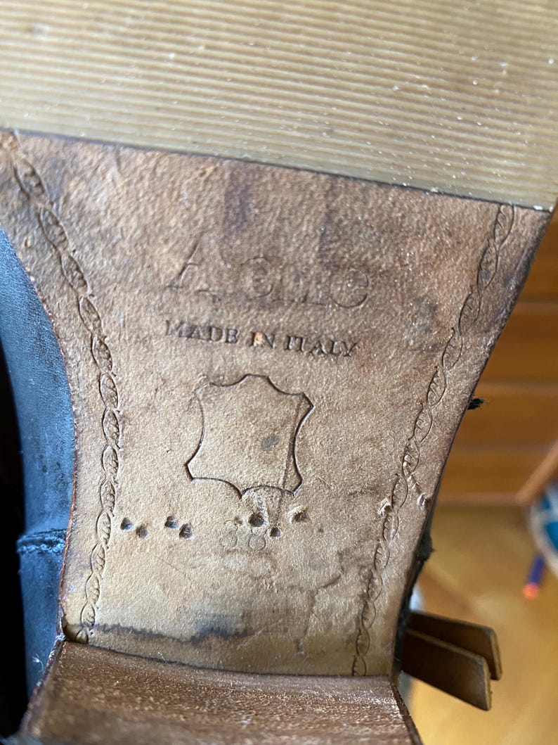 May include: Close-up of the sole of a black leather boot. The sole is made of brown leather and features the text "ACIO MADE IN ITALY". The leather is worn and shows signs of use.