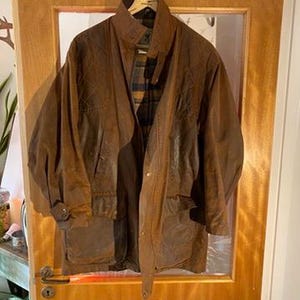 May include: A brown waxed cotton jacket with a high collar and multiple pockets hangs on a wooden hanger. The jacket has a button closure and a plaid lining visible at the chest. The jacket is displayed on a wooden door.
