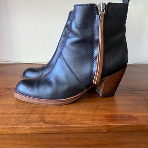 May include: Black leather ankle boots with a wooden heel and a side zipper. The boots have a rounded toe and a pull tab at the back. The heel is approximately 3 inches high.