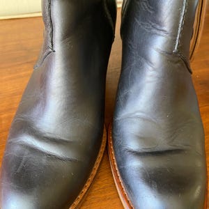 May include: A pair of black leather boots with a classic design. The boots have a smooth, polished finish and a light brown sole. The stitching is visible along the seams, and the boots appear to be well-worn.