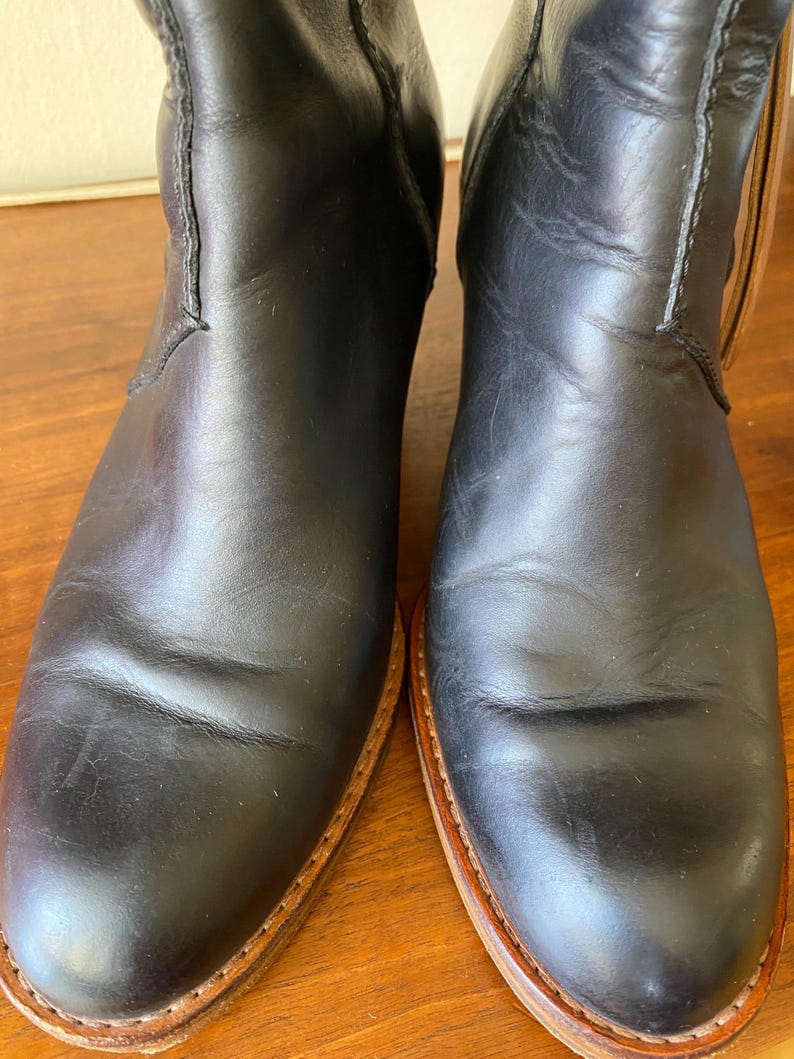 May include: A pair of black leather boots with a classic design. The boots have a smooth, polished finish and a light brown sole. The stitching is visible along the seams, and the boots appear to be well-worn.