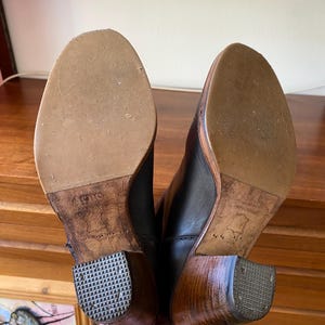 May include: A pair of black leather boots with wooden heels. The soles are light brown with a textured pattern. The heels are a dark brown color with a square, textured bottom. The boots are held up.