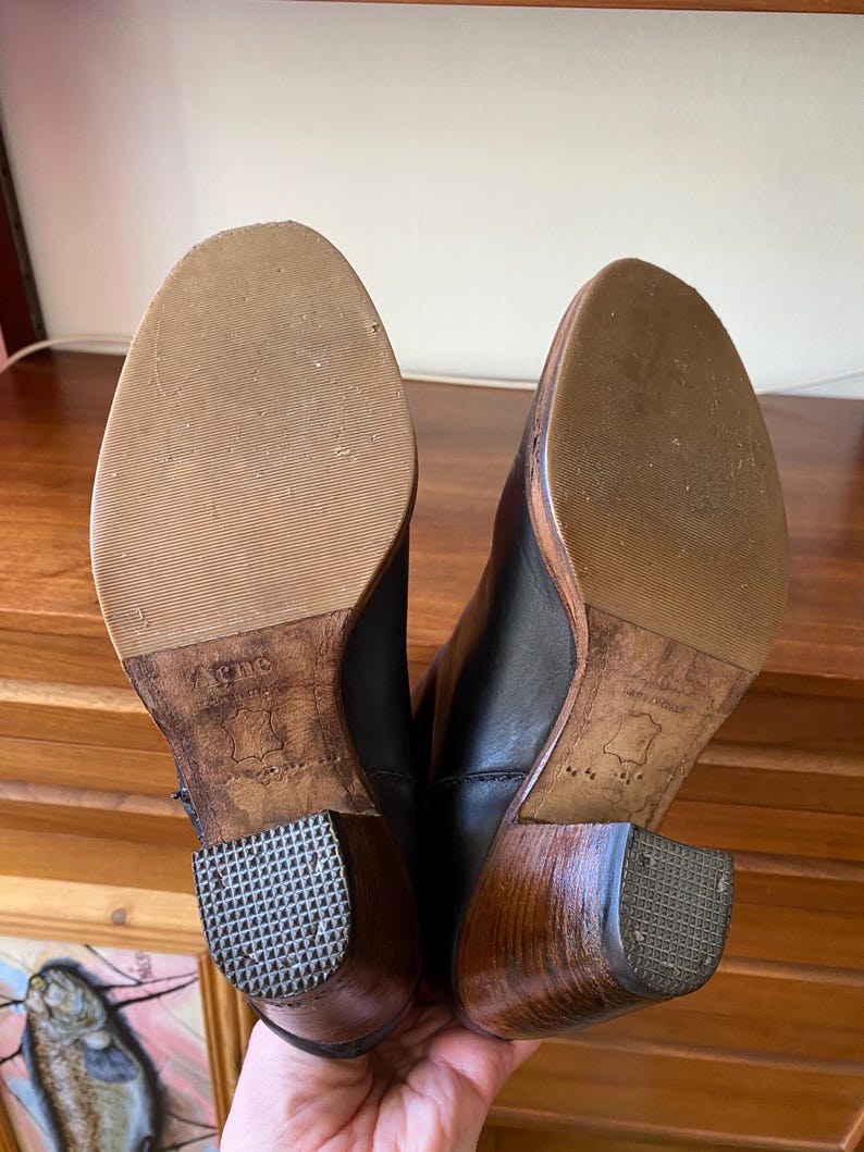 May include: A pair of black leather boots with wooden heels. The soles are light brown with a textured pattern. The heels are a dark brown color with a square, textured bottom. The boots are held up.
