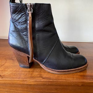 May include: Black leather ankle boots with a wooden heel. The boots have a side zipper with a brown leather pull tab. The sole is a light brown color. The heel is approximately 3 inches high.