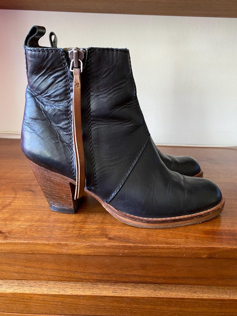 May include: Black leather ankle boots with a wooden heel. The boots have a side zipper with a brown leather pull tab. The sole is a light brown color. The heel is approximately 3 inches high.