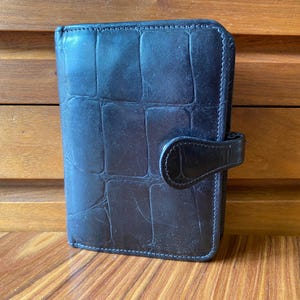 May include: A black leather personal organizer with a rectangular embossed crocodile skin pattern. The organizer features a vertical orientation, stitched edges, and a small side tab closure that secures the front cover to the main body.