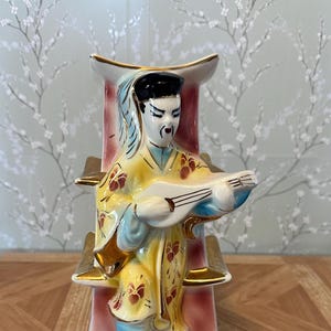 May include: Vintage ceramic figurine of a musician in traditional attire, playing a stringed instrument. The figure is adorned in yellow and blue robes with floral accents, standing on a cloud-like base. The vase has a pink and gold frame.
