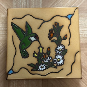 May include: Square ceramic tile featuring a green hummingbird with a long beak, feeding from a flower. The tile has a tan background with black outlines and accents of white, orange, and blue. Decorative tile.