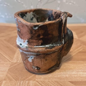 May include: Handmade ceramic mug with a unique, textured design. The mug features a brown and black color scheme with white accents. It has a distinctive handle and a rustic, artisanal aesthetic, perfect for coffee or tea.