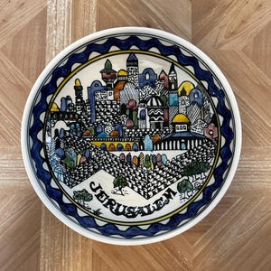 May include: A decorative ceramic bowl with a detailed illustration of Jerusalem. The bowl features a blue and white border, a yellow inner ring, and the word "Jerusalem" in black. The artwork depicts buildings in various colors.