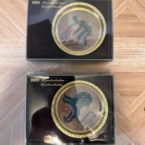 May include: Two framed needlepoint artworks. Each artwork is round with a gold-colored decorative frame. One features a figure in dark blue, the other an octopus. The text "ROPI Handarbeiten - Kostbarkeiten" is printed on the black box.