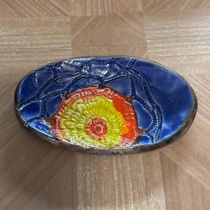 May include: Oval-shaped ceramic dish with a vibrant design. The dish features a deep blue background with a textured, swirling pattern in the center, transitioning from yellow to orange. The rim has a metallic finish.