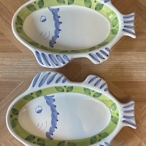 May include: Two white ceramic fish-shaped serving platters. Each platter has a green and blue painted border, with a blue fish design on the side. The platters are oval-shaped and rest on a wooden surface.