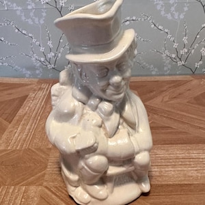 Könnte beinhalten: Cremefarbene Keramikfigur eines Mannes mit Zylinder, der ein kleines Tier hält. Die Figur hat oben eine krugartige Öffnung. Die Figur ist detailliert mit Gesichtszügen und Kleidung, vor einem floralen Tapetenhintergrund.