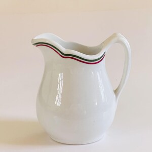 May include: White ceramic pitcher with a wavy rim. The rim is decorated with a thin band of red, green, and red stripes. The pitcher has a curved handle and a rounded body.