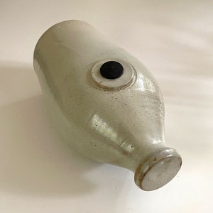 May include: A light gray ceramic bottle with a rounded body and a small, circular base. A black, circular stopper is set into the side of the bottle. The bottle has a speckled texture and a slightly aged appearance.