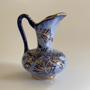 May include: A light blue ceramic pitcher with a dark blue handle and floral design. The pitcher has gold trim around the rim and base. The floral design is in gold. The pitcher is a decorative item.