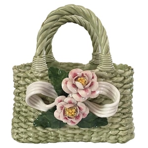 May include: A ceramic basket-shaped decorative piece in a light green hue, featuring a woven texture and a braided handle. It is adorned with two pink and white floral accents and a white ribbon. The overall design evokes a sense of spring and elegance.