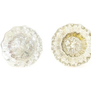 May include: Two clear, vintage glass doorknobs with a floral design. The knobs have a round shape with a scalloped edge and a starburst pattern in the center. The glass has a slight yellow tint.