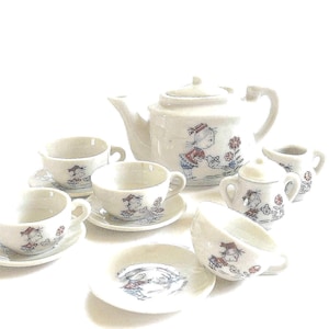 May include: A miniature ceramic tea set featuring a teapot, four cups, four saucers, a lidded sugar bowl, and a creamer. Each piece is decorated with a line drawing of a child in a red outfit watering flowers with a blue watering can.