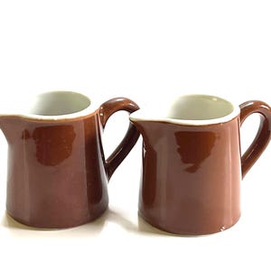 May include: Two small ceramic creamers with a glossy brown exterior finish and a smooth white interior. Each pitcher features a curved handle on the side and a distinct pouring spout, showcasing a simple, functional design for serving liquids.
