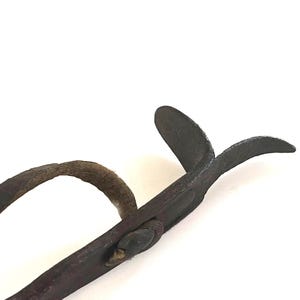 May include: Close-up of a vintage, dark metal tool with a curved handle and scissor-like blades. The tool appears to be made of iron or steel, with a weathered, aged appearance. The handle is curved and the blades are slightly open.