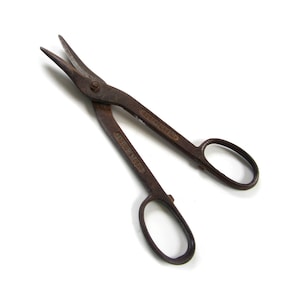May include: Vintage metal shears with a rusty brown finish. The shears have a curved blade and oval finger holes. The text "SOLID STEEL" and "LONDON FORGE BROS" is visible on the handles. The shears are designed for cutting metal.