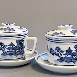 May include: Two white porcelain tea cups with lids and saucers. Each cup features a blue and white Asian-inspired landscape design. The lids have a floral-shaped handle. The cups have a handle and sit on a matching saucer.
