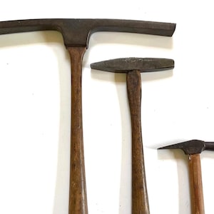 May include: Three vintage hammers with wooden handles and dark metal heads. The hammers vary in size, with the largest featuring a wide, flat head. The handles are a warm brown color, and the metal shows signs of age.