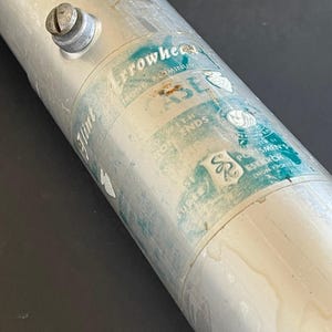 May include: Close-up of a cylindrical metal object with a screw on the side. The object has a label with the text "Arrowhead Case" and other faded text. The object is silver with some wear and tear.