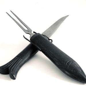 May include: A black carving set featuring a knife and a fork. The handles are textured and shaped like fish. The fork has two tines. The knife blade is silver. The set is crossed over a white background.