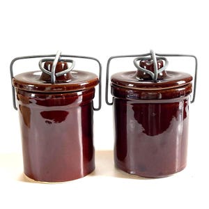 May include: Two matching brown ceramic jars with lids and metal handles. The jars have a glossy finish and a cylindrical shape. The lids are rounded with a small knob. The metal handles are attached to the lids and the sides of the jars.