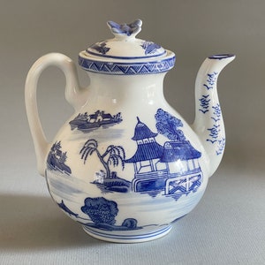 May include: White ceramic teapot with a blue and white landscape design. The teapot features a curved handle, a spout, and a lid with a small bird-shaped handle. The design includes pagodas, trees, and boats, creating a classic, vintage aesthetic.