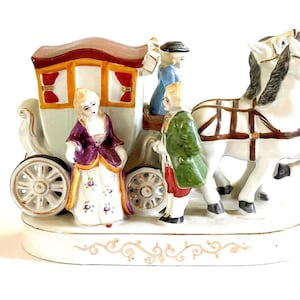 May include: A porcelain figurine depicting a horse-drawn carriage with three figures. The carriage is light green with red and gold accents. The horses are white, and the figures are dressed in period clothing. The base is white with gold scrollwork.