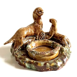May include: A glazed ceramic figurine featuring two brown otters positioned on a textured, multi-colored base with a central circular bowl. The otters have detailed fur textures and are arranged around the small, round, recessed dish.