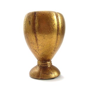May include: A small, goblet-shaped decorative object with a metallic gold finish. The surface features a textured, leaf-like appearance with vertical ridges running along the rounded body, tapering down to a narrow neck and a circular pedestal base.
