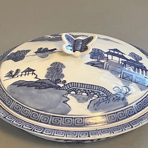 May include: Oval ceramic serving dish with a lid, featuring a blue and white landscape design. The lid has a butterfly-shaped handle. The rim is decorated with a geometric pattern. A decorative piece for serving food.