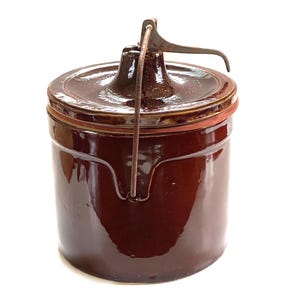 May include: A cylindrical dark brown glazed stoneware crock with a matching lid. The lid features a central knob and a metal wire bail handle that extends over the top. The exterior surface has a smooth, reflective finish with a deep brown color.