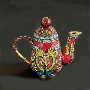 May include: A small, decorative teapot with a red base and lid, adorned with floral patterns in green, blue, and gold. The teapot features a curved handle and spout, with gold accents. The overall design is intricate and ornate.