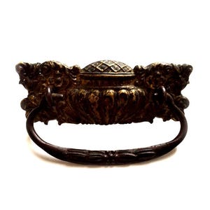 May include: Ornate antique bronze drawer pull with a decorative handle. The pull features detailed floral and scrollwork designs, with a central, textured dome. The handle is curved and has a decorative pattern.