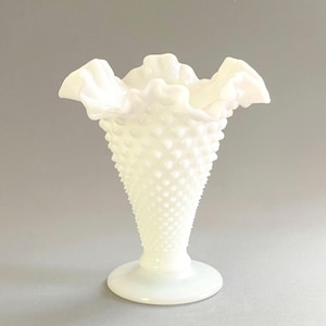 May include: A white hobnail glass vase with a ruffled rim. The vase has a flared shape and a round base. The hobnail texture covers the body of the vase. The vase is likely vintage and suitable for displaying flowers or as a decorative piece.