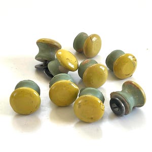 May include: A collection of vintage cabinet knobs. The knobs feature a round, yellow top and a green base. Some knobs are stacked together, while others are scattered on a white surface. The knobs have a distressed, antique appearance.
