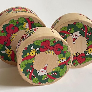 May include: Three wooden Christmas storage boxes. Each box features a festive wreath design with red bows, green holly, and cartoon characters. The boxes are cylindrical and vary in size, perfect for holiday decor and storage.