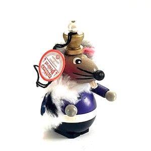 May include: A wooden mouse figurine wearing a gold crown, purple jacket, and black pants. The mouse has a red nose and a white fur collar. A red tag with the text "Steinbach Handmade Germany" hangs from the figurine.