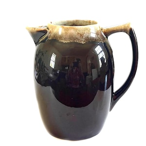 May include: A glossy, dark brown ceramic pitcher with a curved handle and a spout. The top rim is a lighter tan color. The pitcher reflects the surrounding environment, creating a mirrored effect. A vintage kitchenware item.